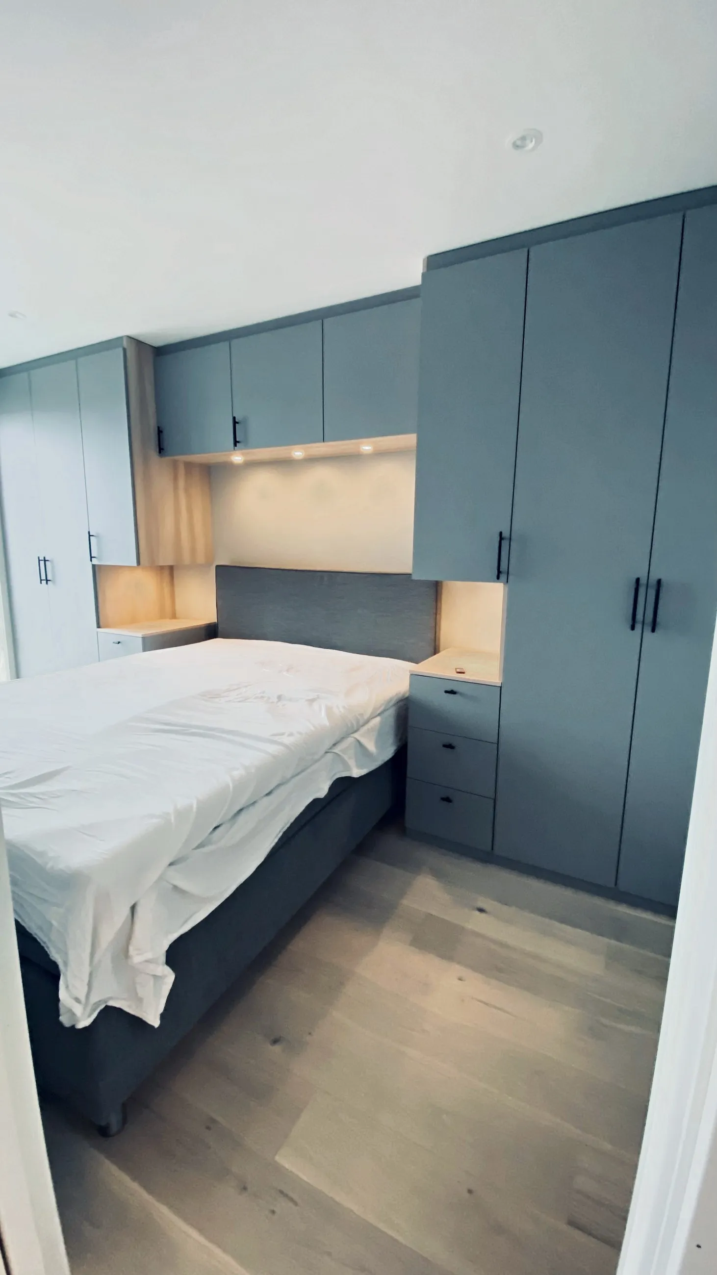 Garderobeskap bygget rundt sengen i blått, med overskap, nattbord og LED-lys under skapene