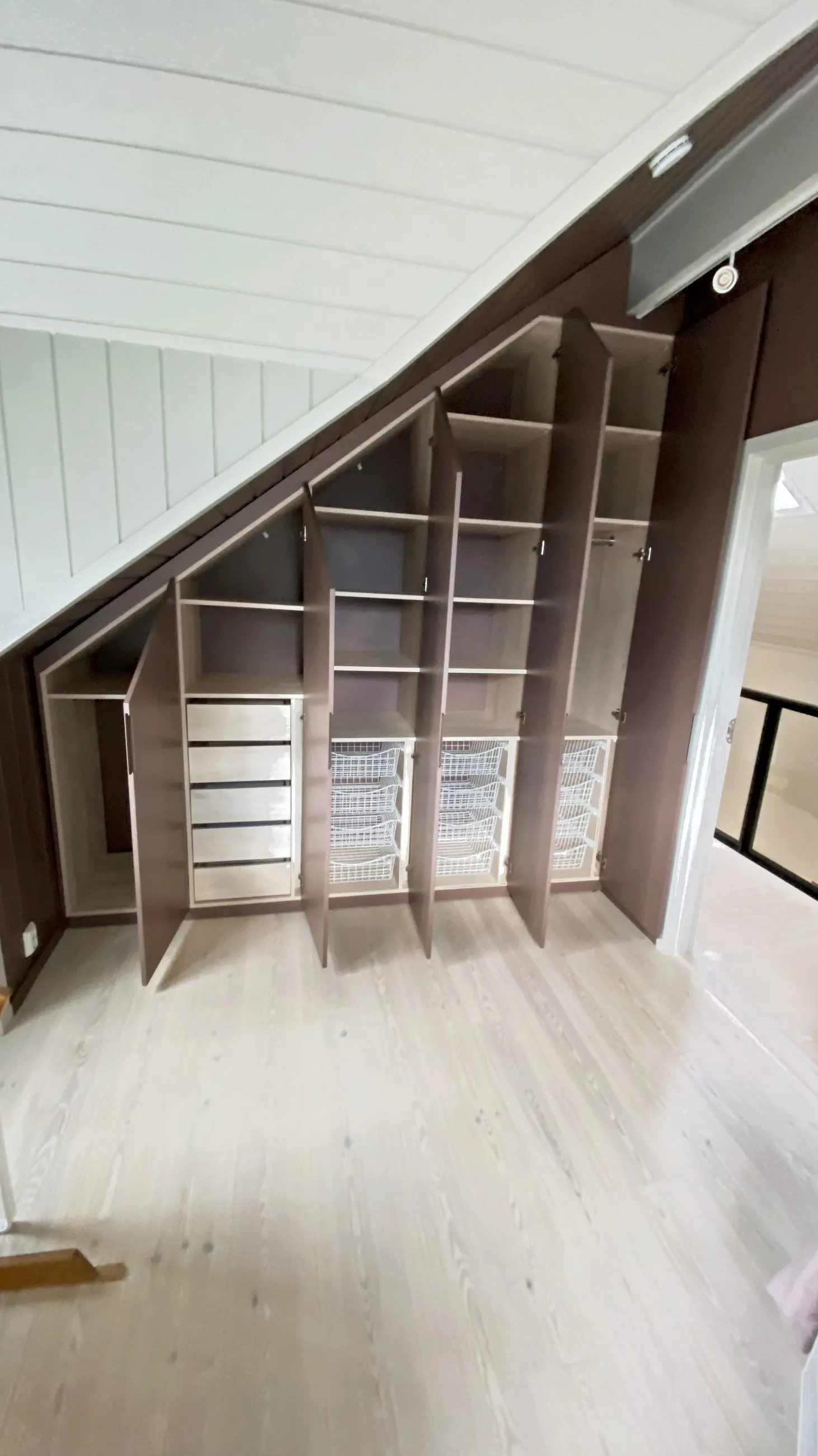 Mørkebrun skråtaksgarderobe med åpne dører, og hyller, skuffer og trådkurver innenfor